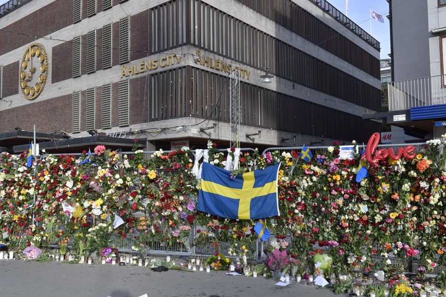 epa05898352 A Swedish flag, floral tributes and candles on a fence by the department store, Ahlens, beside the scene of a truck attack in Stockholm, Sweden, 09 April 2017. The flowers are being moved  ...