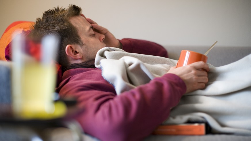 [Themenbild Grippe, gestellte Aufnahme] Ein Mann, der erkrankt ist, liegt mit einer Tasse Tee auf dem Sofa und haelt sich den schmerzenden Kopf, aufgenommen am 27. Oktober 2007 in Zuerich. (KEYSTONE/M ...