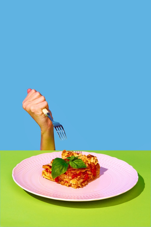 Female hand holding fork over delicious Italian food, lasagna over green and blue background. Bolognese sauce and basil. Concept of Italian food, cuisine, taste, cooking, menu. Pop art. Poster, ad