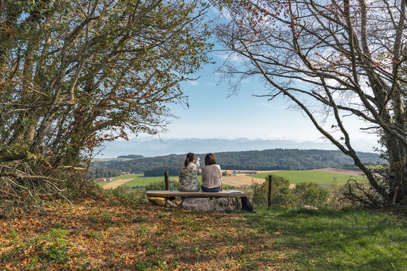 Zwei Freundinnen sitzen auf einer Waldlichtung auf einer Bank und geniessen die Aussicht auf den Genfersee und die Berge dahinter.