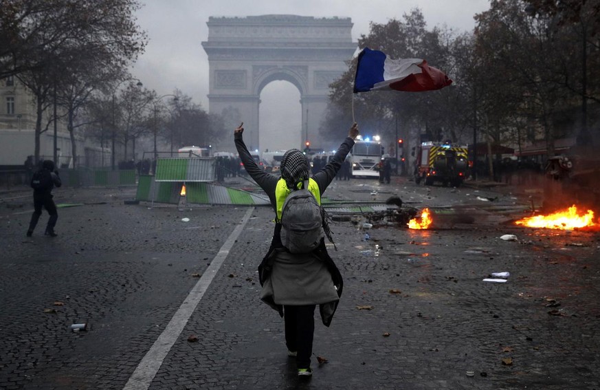 epa07201551 A protester wearing a yellow vest (gilets jaunes) waves a French flag during clashes with riot police near the Arc de Triomphe as part of a demonstration over high fuel prices on the Champ ...