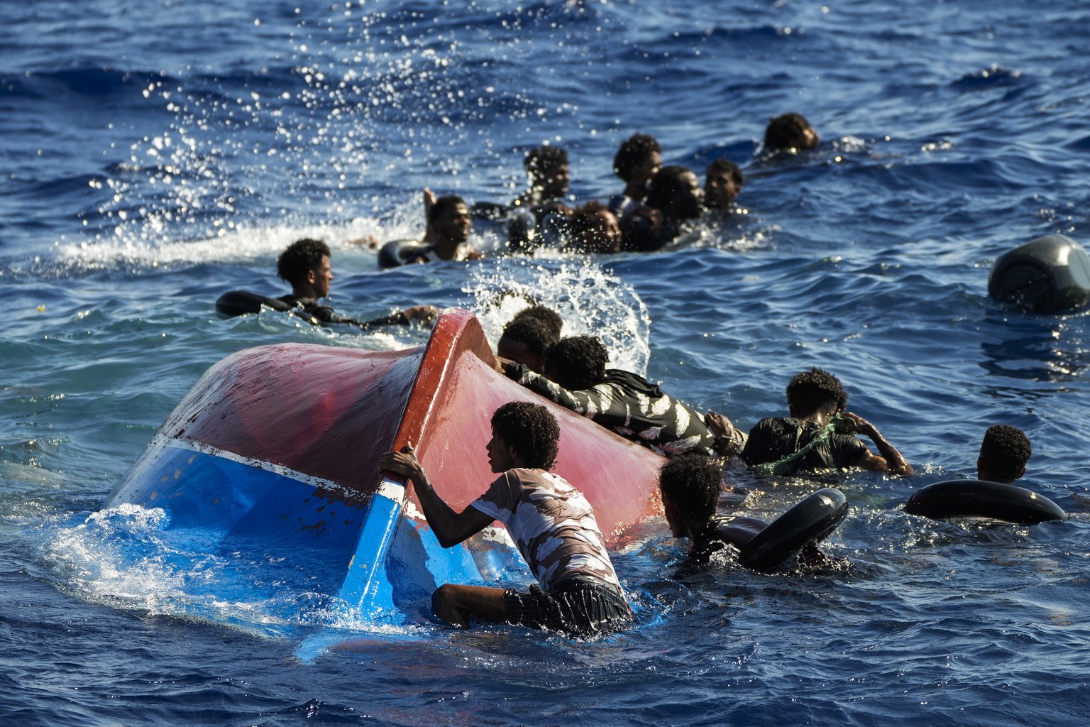 FILE - Migrants swim next to their overturned wooden boat during a rescue operation by Spanish NGO Open Arms at south of the Italian Lampedusa island at the Mediterranean sea, Aug. 11, 2022. The back- ...