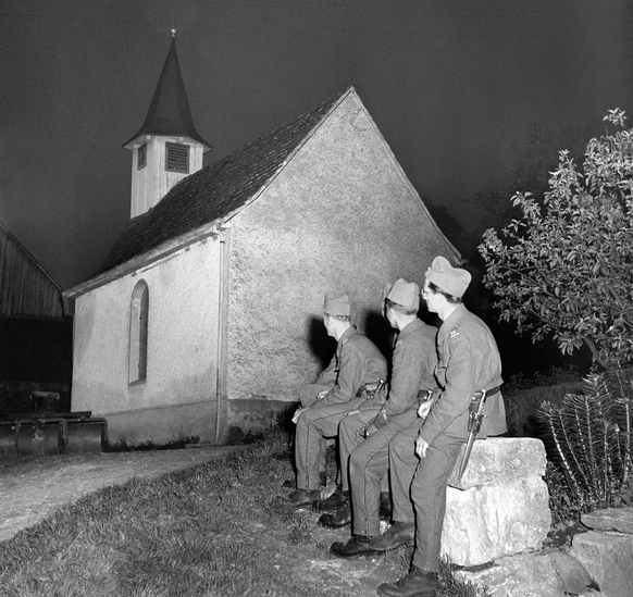 Devant une petite église du canton d'Argovie, des soldats écoutent la sonnerie des cloches qui retentit pendant un quart d'heure dans tout le pays pour célébrer la fin de la Seconde Guerre mondiale.