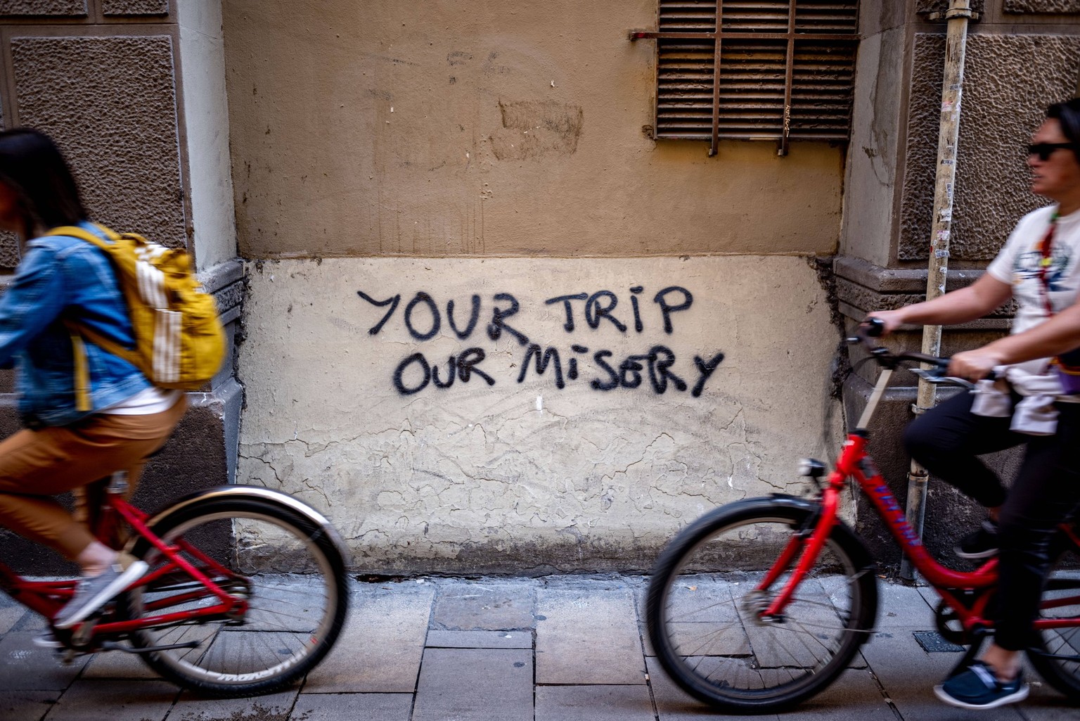 April 2, 2024, Barcelona, Spain: In Barcelona s Gothic Quarter a group of tourists on a bike tour pass an anti-tourism graffiti reading Your Trip Our Misery. Protests against mass tourism have been go ...