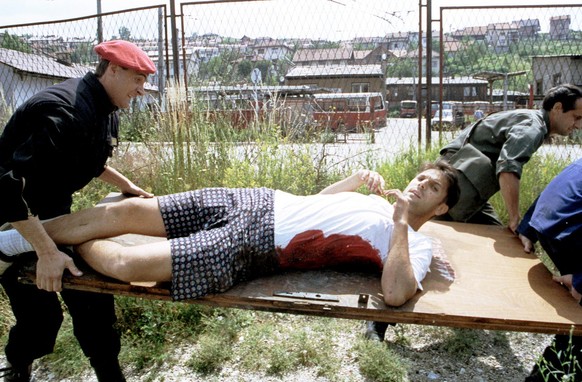 Le 17 juin 1993, Des civils de Sarajevo, aidés par un soldat isolé, transportent un jeune homme blessé par un tireur embusqué vers un lieu sûr en utilisant une porte comme civière.