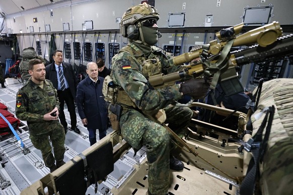 epa10933912 German Chancellor Olaf Scholz (3-L) stands behind a soldier with a machine gun on top of a buggy of the air mobile protection team during a flight in an A400M Airbus from BER government te ...