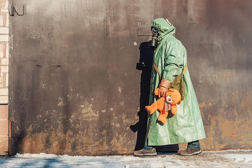 Un homme en combinaison radioprotection et masque à gaz portant un ours en jouet dans la rue de la ville couverte de neige. Concept de l&#039;ère nucléaire.