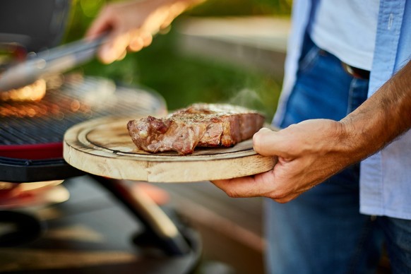 Man put down on a wooden board ready to eat grilled steak meat, male roasted steak meat on the gas grill on barbecue grill outdoor in the backyard, summer family picnic, food on the nature.