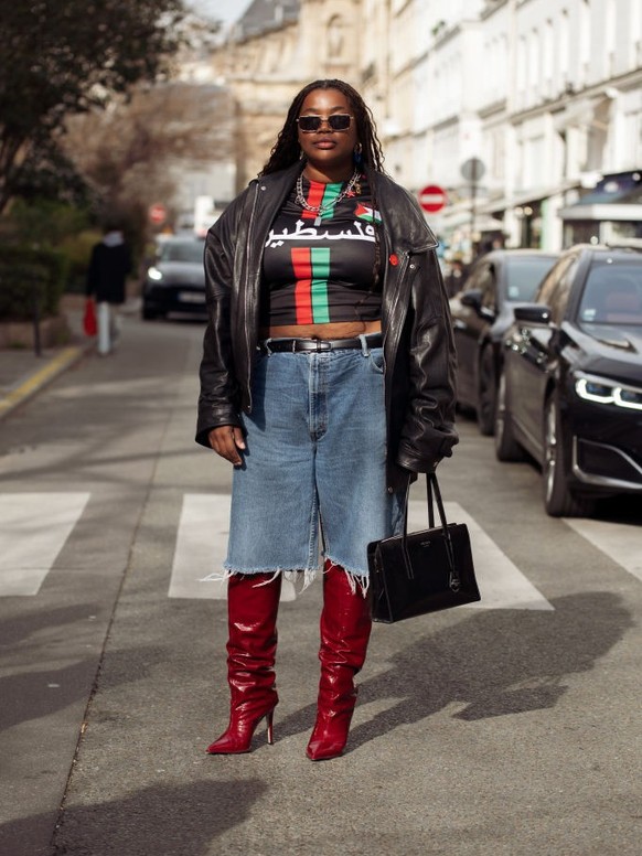 PARIS, FRANCE - FEBRUARY 28: Gabriella Karefa-Johnson wears dark deep red boots, blue denim shorts Bermuda, black red and green shirt, black leather jacket and sunglasses outside The Row during the Wo ...