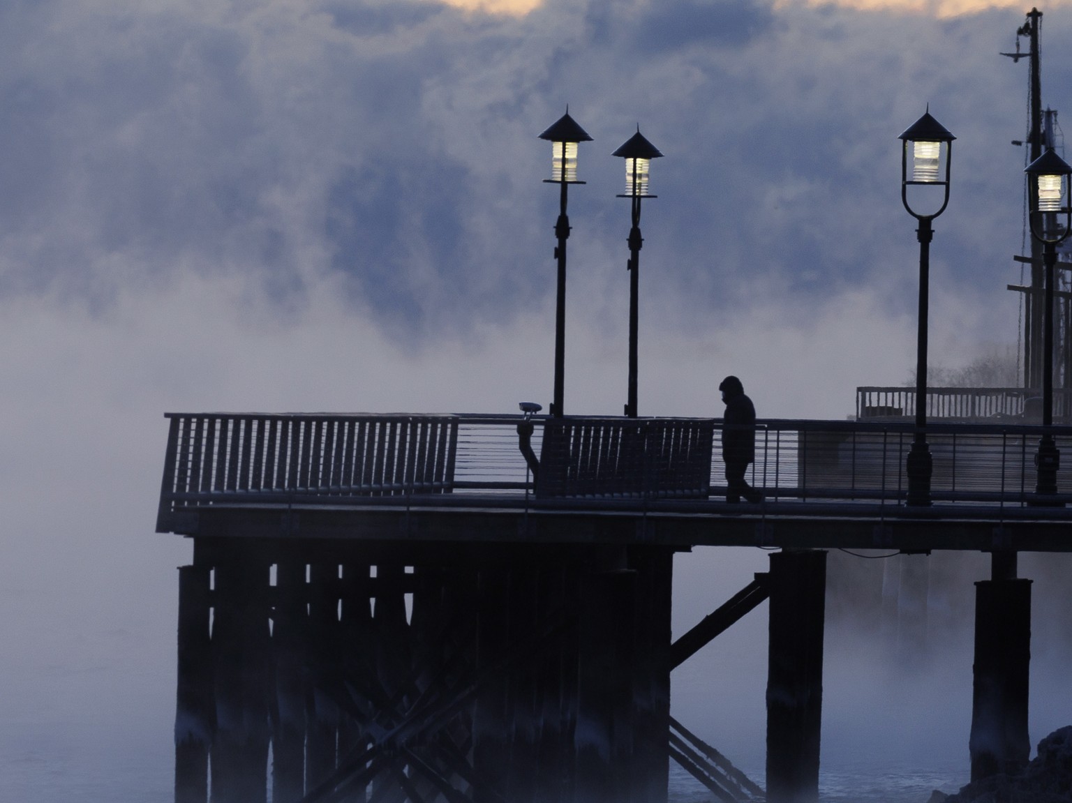 epa10447086 A person walks on a pier as sea smoke forms over the water of Boston Harbor, in Boston, Massachusetts, USA, 04 February 2023. Sea smoke, or steam fog, forms when cold air flows immediately ...
