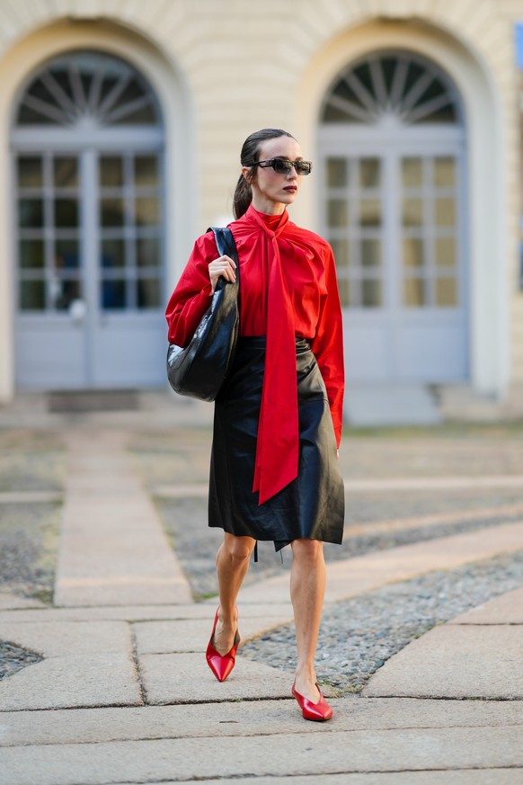 MILAN, ITALY - SEPTEMBER 24: A guest wears black sunglasses, a red oversized wrap shirt with a knotted neck, a black shiny leather shoulder bag, a black shiny leather high waist knees skirt, silver ri ...