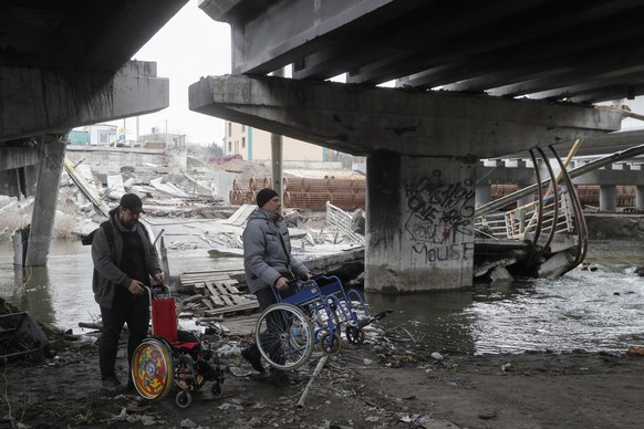 Des hommes transportent des fauteuils roulants sous le pont Romanov détruit entre Irpin et Kiev en Ukraine, 31 mars 2023.