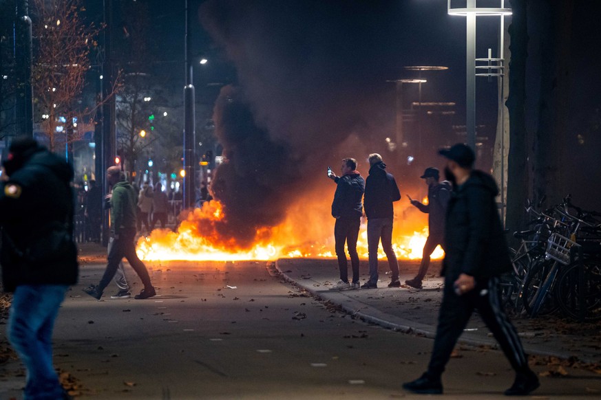 epa09593018 People stand near burning objects after a protest against the &#039;2G policy&#039; turned into riots, with protesters setting fires in the street and destroying police cars and street fur ...