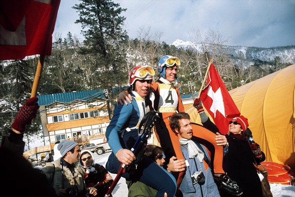 Der Schweizer Abfahrts-Olympiasieger Bernhard Russi (Nr. 4) und der Silbermedaillengewinner Roland Collombin (links) auf den Schultern von Fans an den Olympischen Winterspielen in Sapporo 1972. Die &# ...