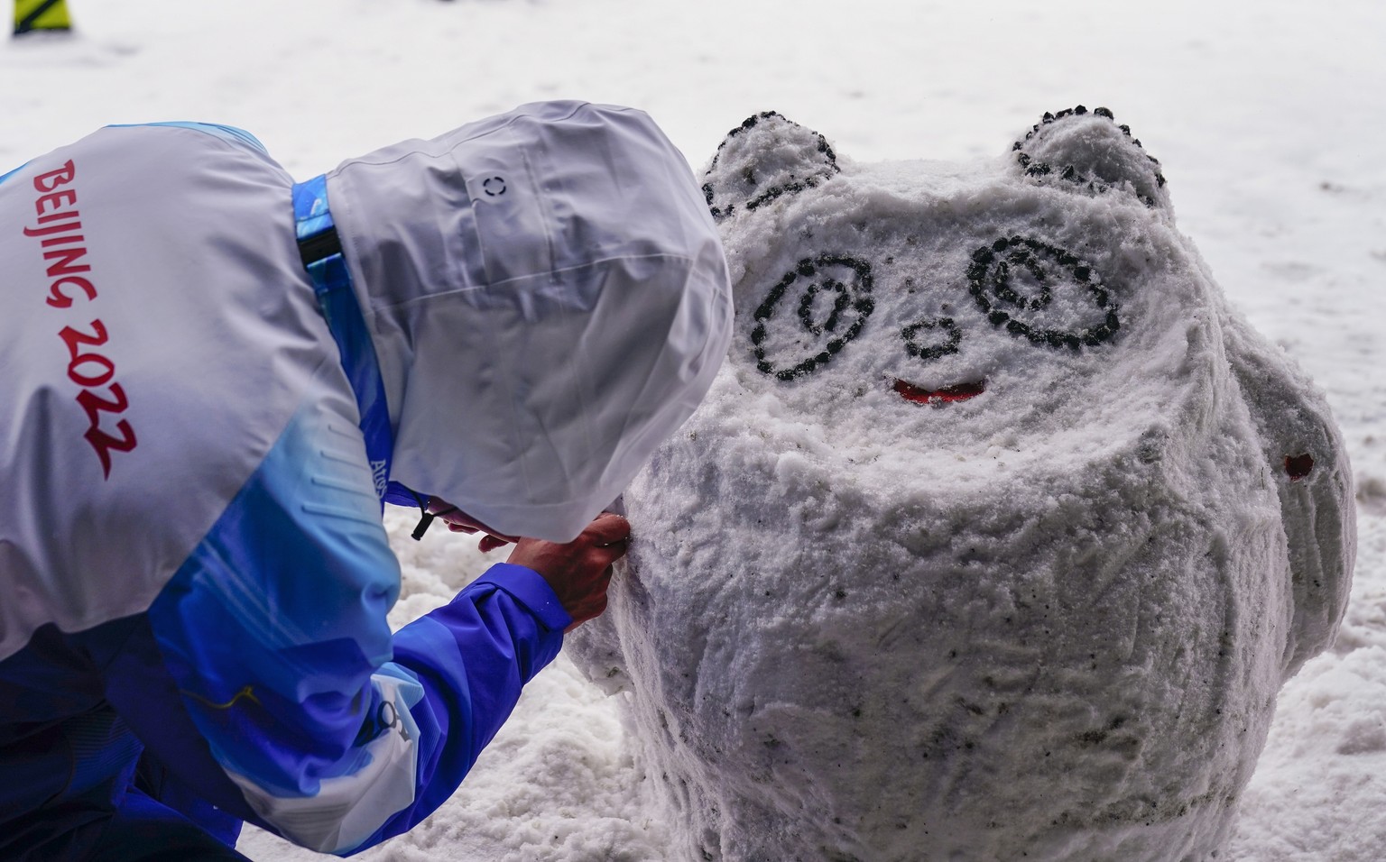 A volunteer makes a snow version of the olympic mascot Bing Dwen Dwen near the Zhangjiakou National Ski Jumping Centre at the 2022 Winter Olympics, Sunday, Feb. 13, 2022, in Zhangjiakou, China. (AP Ph ...
