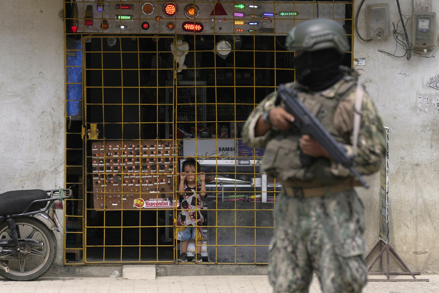 A boy watches from behind a storefront gate as a soldier stands guard at a security check point, in Duran, Ecuador, Monday, Aug. 14, 2023. Ecuador&#039;s president declared a state of emergency in som ...