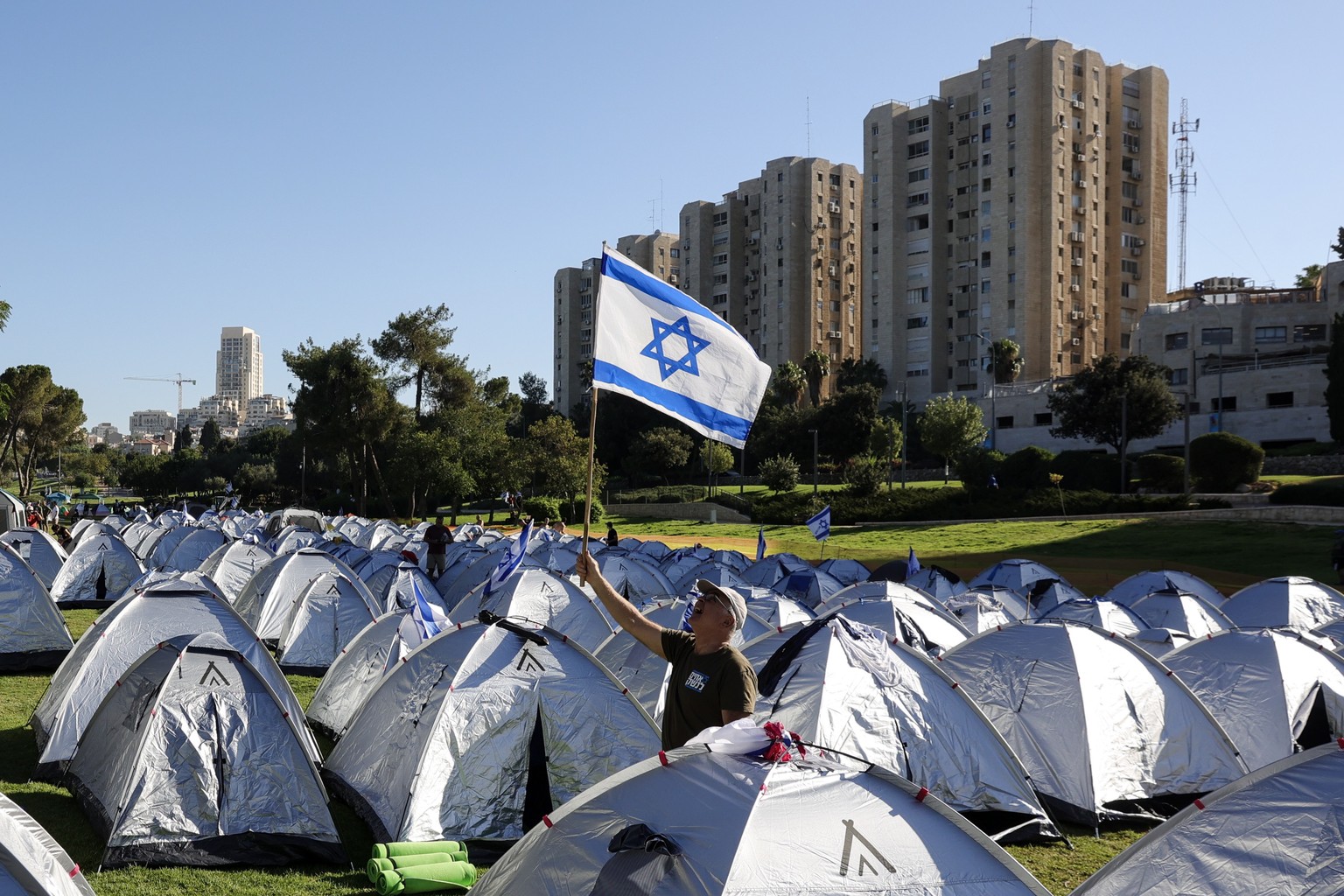 epa10763324 An anti-government raises a Israeli flag among tents after spending the night in a tent camp at Sacher Park, near the Israeli Knesset, following a four-day protest march to Jerusalem again ...