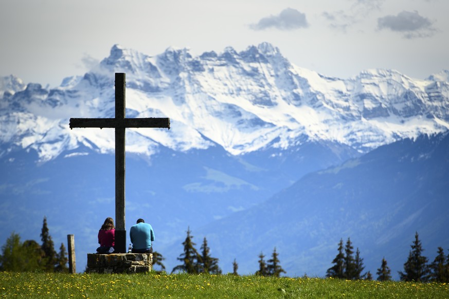People take a break under a cross in a field and enjoy a spring weather day in front of the Swiss Alps mountains (Les Dents du Midi), in Les Pleiades Blonay above Montreux, Switzerland, Sunday, May 23 ...