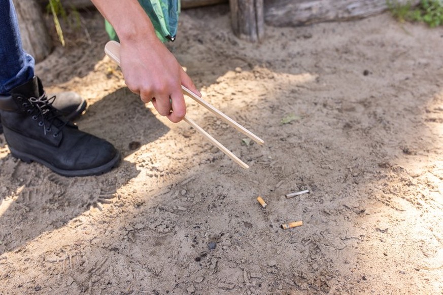 Hand of a volunteer picking up cigarette buds using a wooden tong. Close-up of a person collecting the trash from the forest.