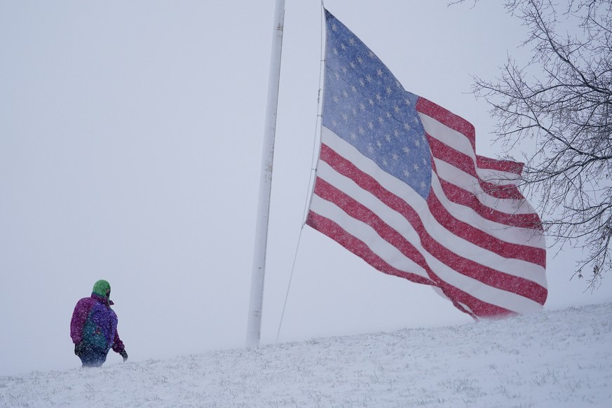A person walks along a path as a large United States flag waves in gusty wind during a snow storm, Monday, Jan. 3, 2022, in Baltimore. (AP Photo/Julio Cortez)