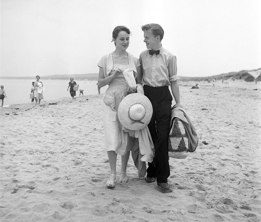 1950er Jahre am Strand Ein junges Paar ist gerade am Strand angekommen und geht auf der Suche nach einem schnen Platz fr einen sonnigen Tag spazieren Sie sind gut gekleidet wie man es in den 1950er Ja ...