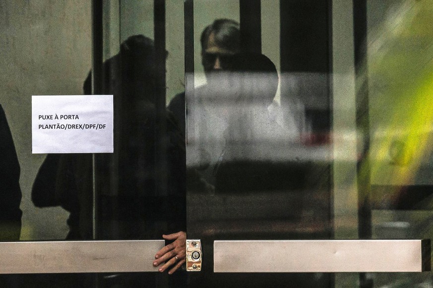 epa12544836 Former Brazilian President Jair Bolsonaro (C) stands behind a glass door at Federal Police headquarters in Brasilia, Brazil, 23 November 2025. At a detention review hearing, Bolsonaro said ...