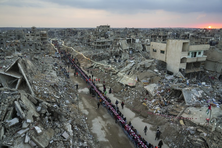 As the sun sets, Palestinians sit at a large table surrounded by the rubble of destroyed homes and buildings as they gather for iftar, the fast-breaking meal, on the first day of Ramadan in Rafah, sou ...