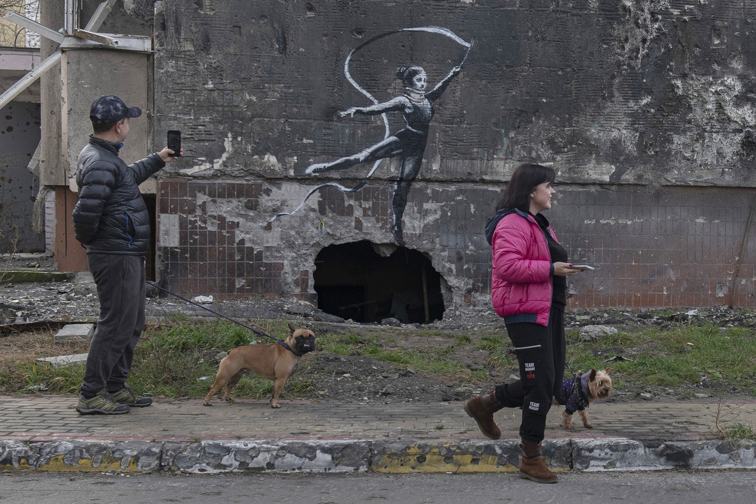 People walk their dogs past an artwork on a building destroyed by fighting in Irpin, Kyiv region, Ukraine, Sunday, Nov. 13, 2022. The painting appears to have been made by British street artist Banksy ...