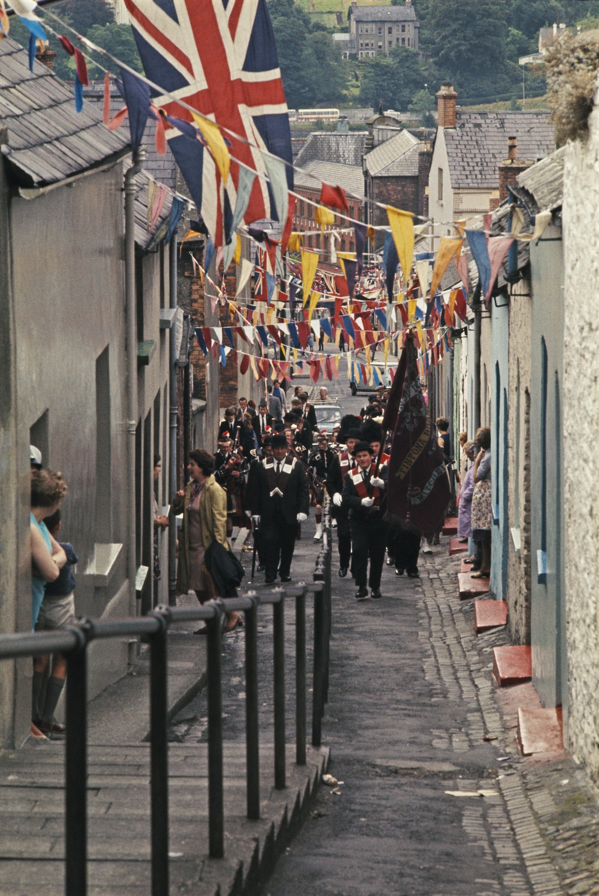 View of an annual Apprentice Boys' parade, with Protestant pipe band members marching and playing along a street decked with bunting and union flags, to celebrate the lifting of the siege of the  ...