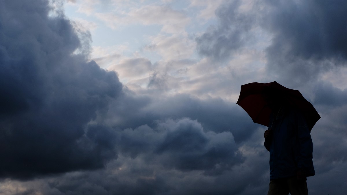 ARCHIV - 12.07.2019, Nordrhein-Westfalen, Duisburg: Ein Mann geht mit einem Regenschirm vor aufziehenden dunklen Wolken spazieren. Erst zu wenig, dann zu viel Regen