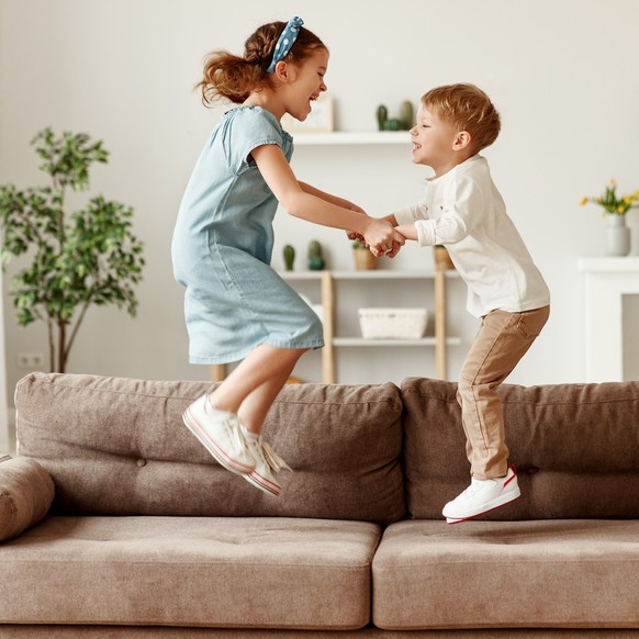 Side view of cheerful boy and girl holding hands and jumping on couch while having fun at home togethe model released