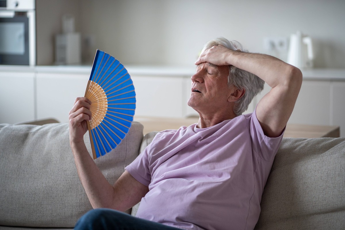 Älterer Mann kämpft mit Hitzeerschöpfung durch Ventilatoren. Rentner versucht, im Sommer in der Hitze des Hauses zurechtzukommen Elderly man feeling dizzy and unwell from heat, fanning himself at home ...
