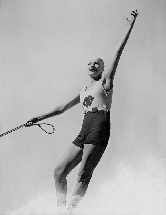 Water-skiing model with one arm raised, the other holding her tether, wearing swim cap and bathing suit: heavy white jersey tank top; high-waisted, short-legged and belted jersey trunks; designed by H ...