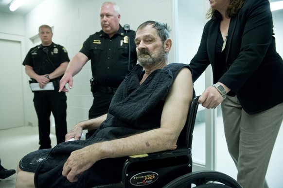 Der Angeklagte wird in einem Rollstuhl zur Videoanhörung geführt.