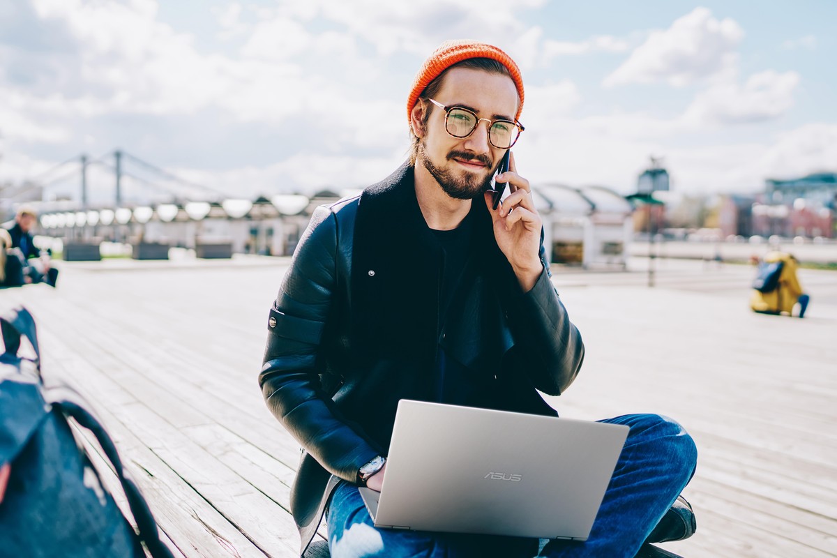 Portrait of smiling hipster guy in trendy wear having mobile phone conversation while sitting outdoors with laptop computer, adult man student making cellphone call in roaming looking at camera; Shutt ...