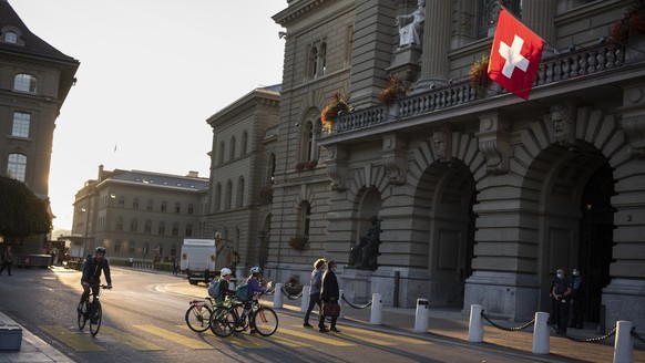 Leute ueberqueren den Fussgaengerstreifen vor dem Bundeshaus, waehrend die Morgensonne die Schweizer Fahne am Parlamentsgaebaeude erhellt, an der Herbstsession der Eidgenoessischen Raete, am Dienstag, ...