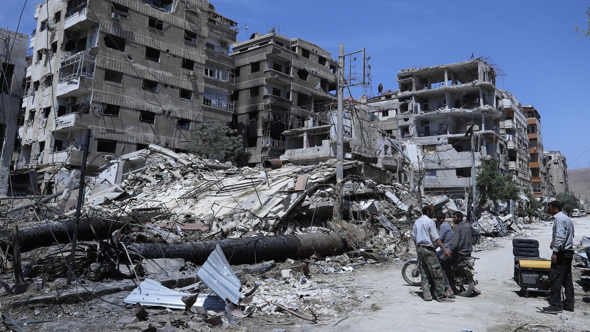 FILE - In this Monday, April 16, 2018 file photo, people stand in front of damaged buildings, in the town of Douma, the site of a suspected chemical weapons attack, near Damascus, Syria. The global ch ...