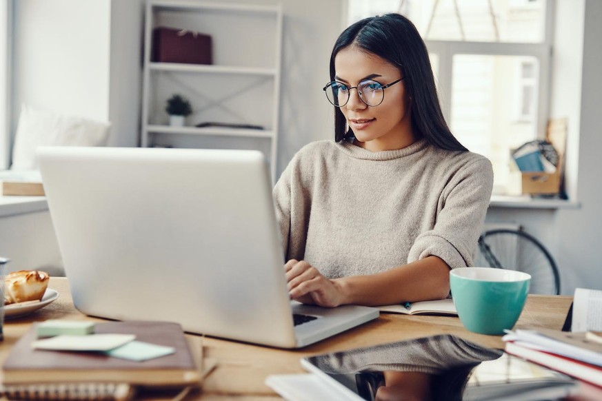 Beautiful young woman in casual clothing using laptop and smiling while working indoors