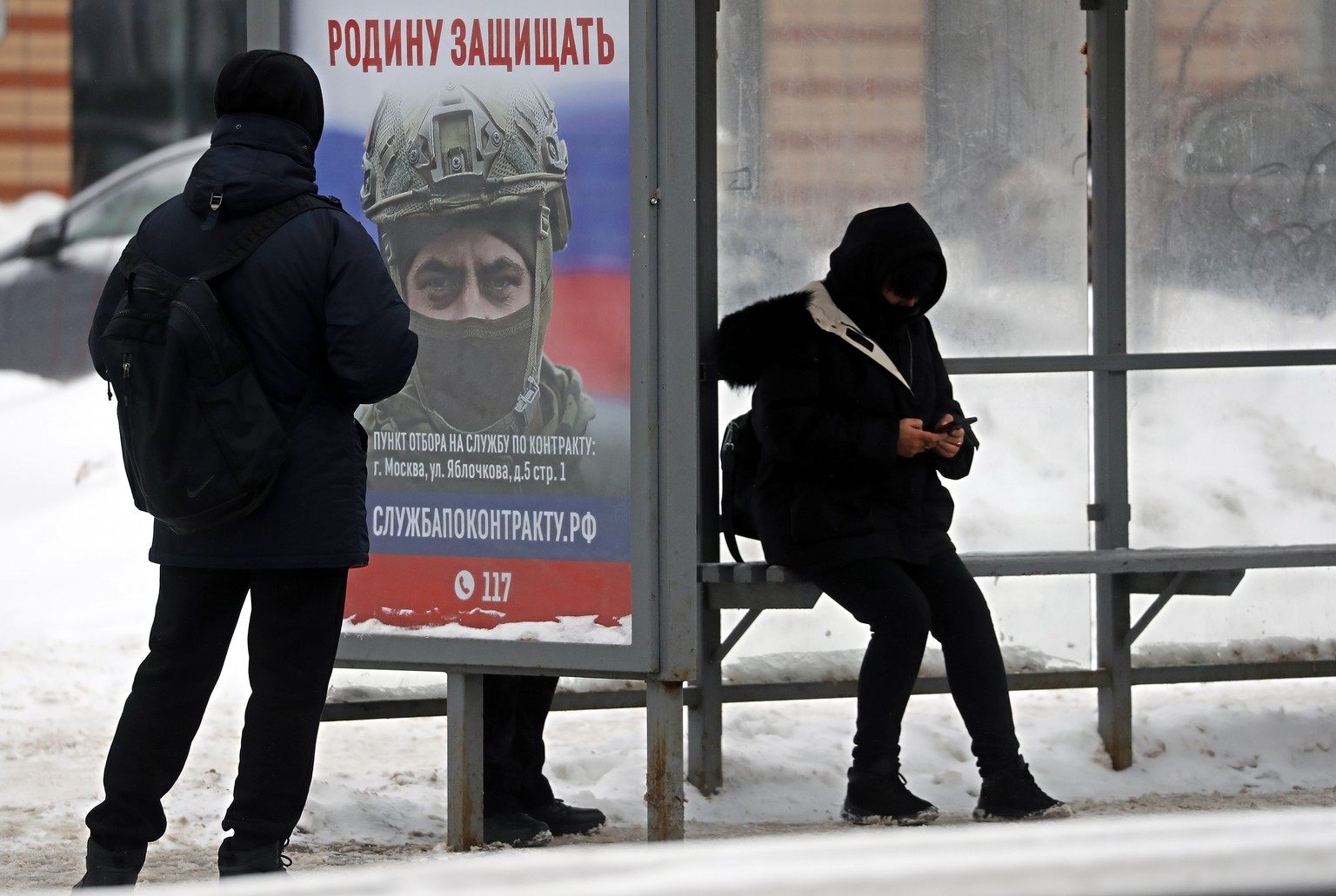 epa11023013 People wait for a bus at a bus stop with advertising military conscription, showing a Russian soldier and text reading &#039;Defend the Motherland. ?ontract service&#039; on a billboard, a ...
