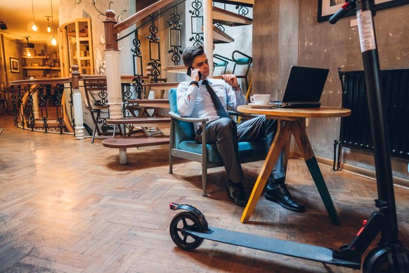 Young caucasian handsome businessman using laptop and talking on mobile phone in cafe and drinking coffee. Electric scooter standing next to him.