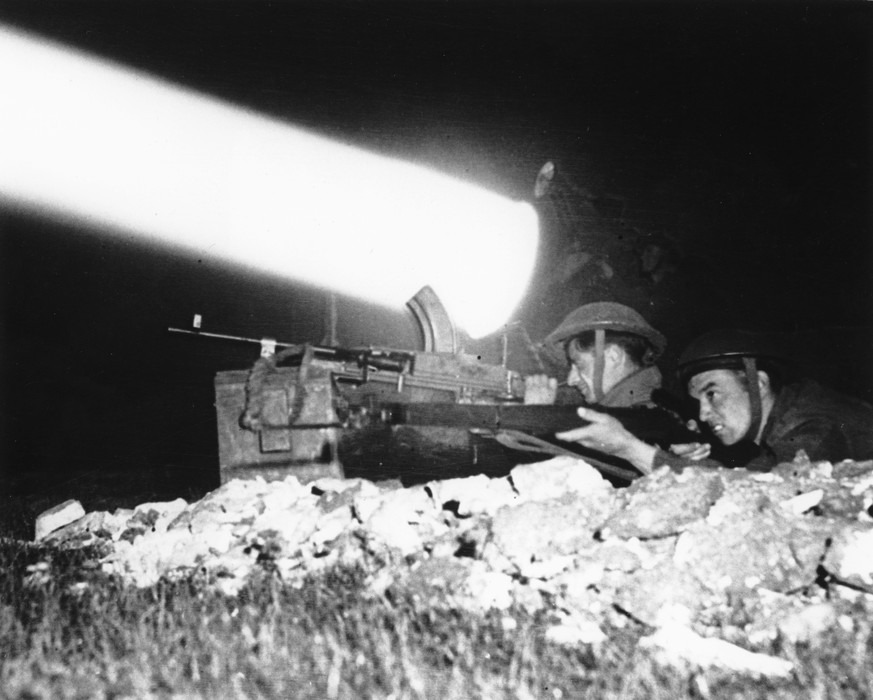 FILE - With a searchlight beam piercing through the night, a machine gunner and a rifleman are on alert as the British Canadian offensive drive continues, in July 1944, south of Caen, in the Normandy  ...