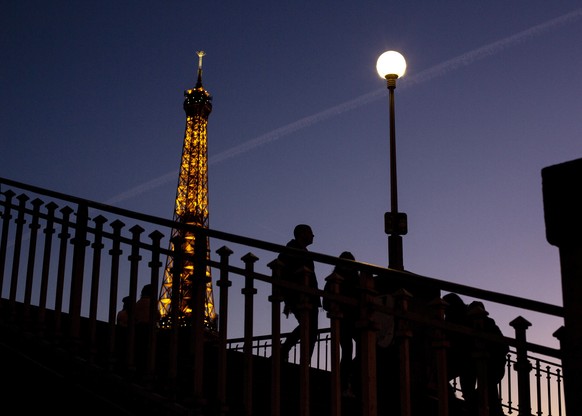 FILE - People walk on a bridge next to the Eiffel Tower in Paris, Wednesday Feb. 9, 2022. Lights on the Eiffel Tower will soon be turned off an hour earlier at night as part of an energy savings plan  ...