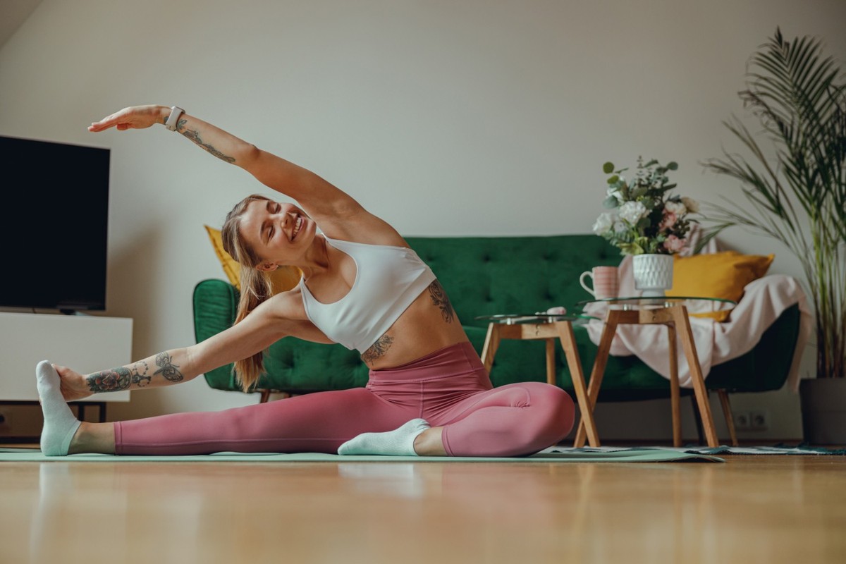 A woman is leisurely stretching her legs on a yoga mat in a living room with hardwood flooring
