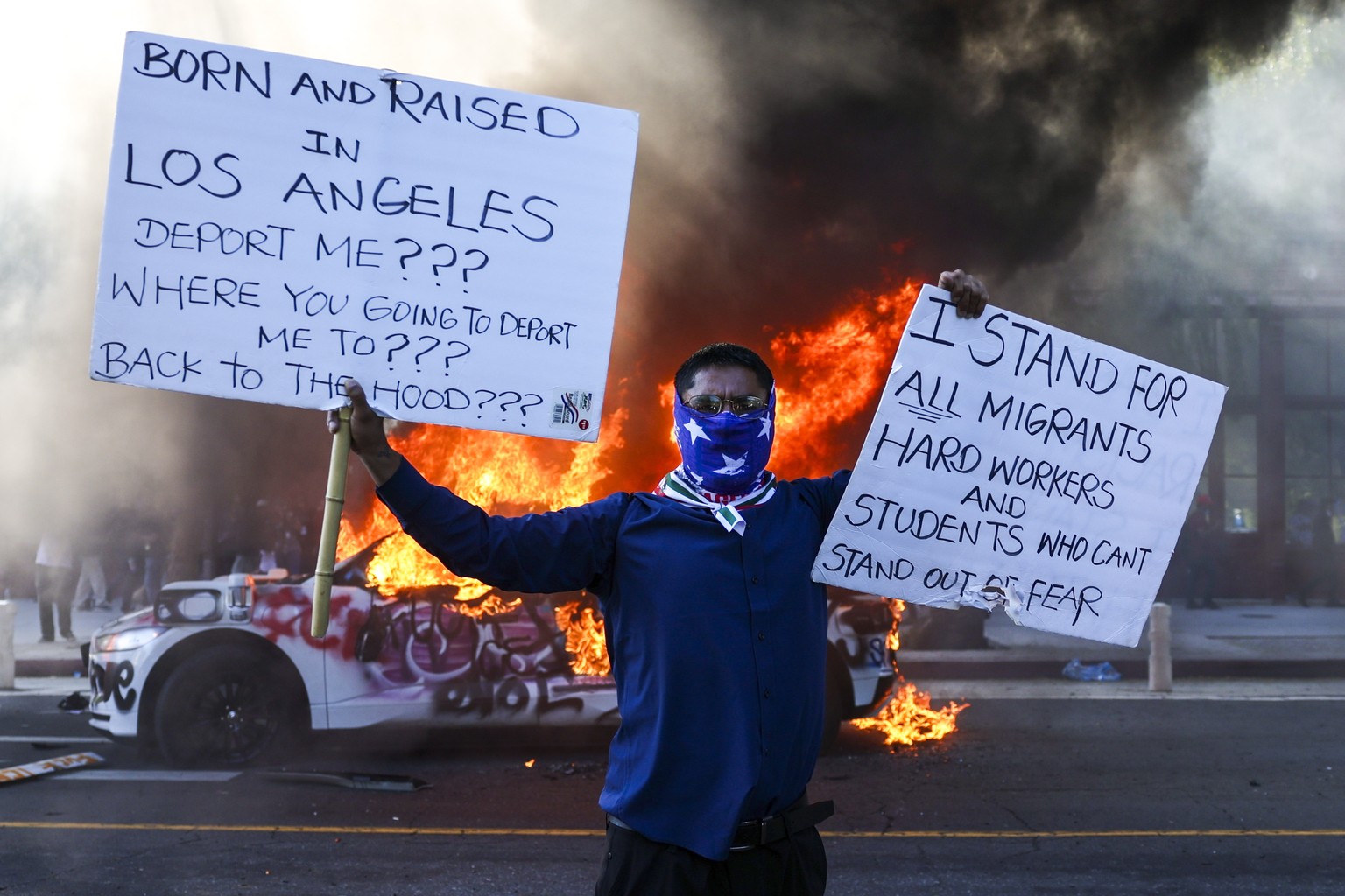 epa12165564 An activist holds signs as a car burns during protests sparked by immigration raids in Los Angeles, California, USA, 08 June 2025. US President Donald Trump has deployed 2,000 National Gua ...