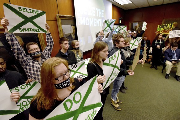 Supporters of victims of sexual abuse by Larry Nassar, a sports medicine doctor for both Michigan State and USA Gymnastics, hold up signs during a Michigan State Board of Trustees meeting, Friday morn ...