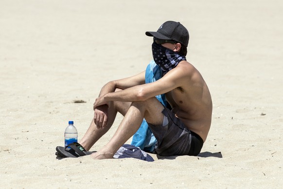 epa08398136 A man wearing a bandana as a face mask sits in the sand on the closed beach amid the coronavirus pandemic in Huntington Beach, California, USA, 02 May 2020. California Governor Gavin Newso ...