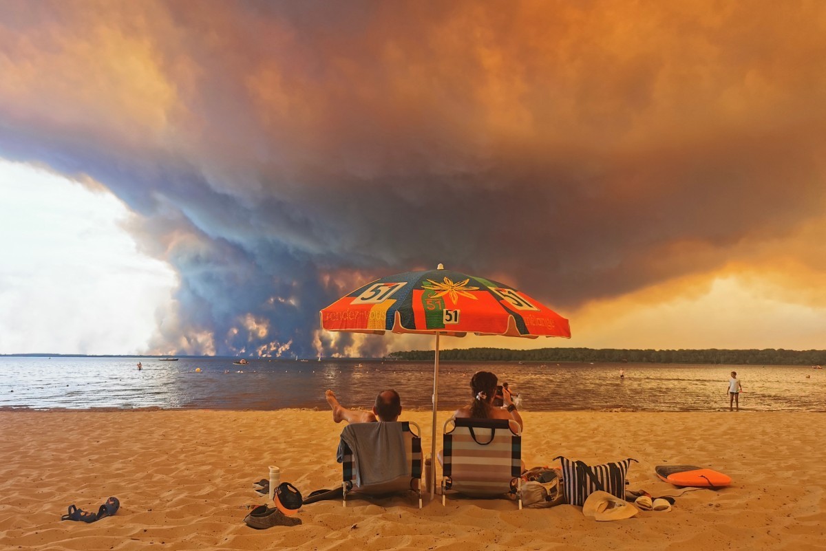 Beach goers sit on the beach of Moutchic in Lacanau, southwestern France on July 24, 2026, as clouds of smoke rise into the sky from a wildfire. The fire raging in the northern part of the Arcachon Ba ...