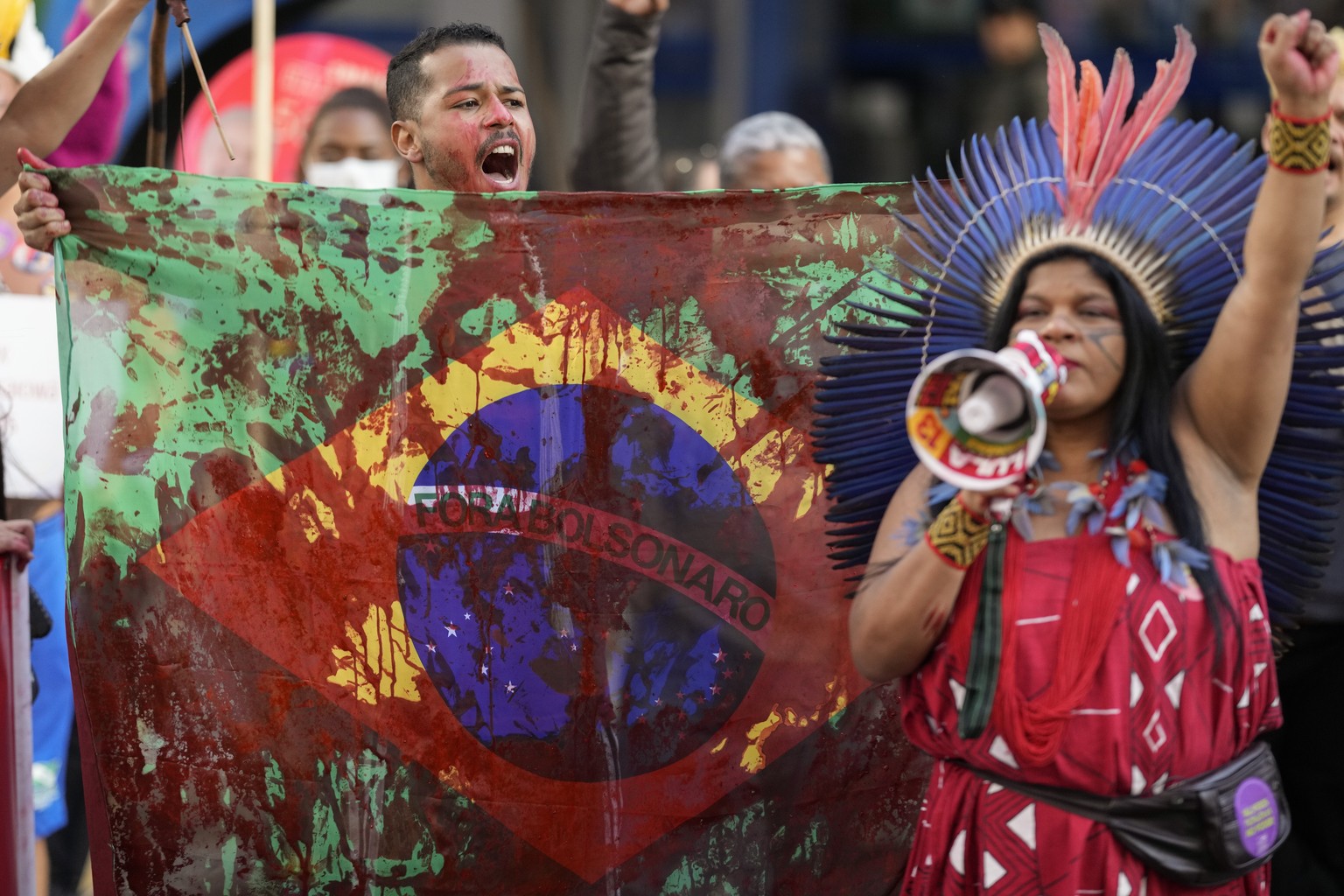 An Indigenous man holds a Brazilian national flag painted in red to symbolize blood, and with the words &quot;Bolsonaro get out&quot; written in Portuguese, during an Indigenous protest against Violen ...