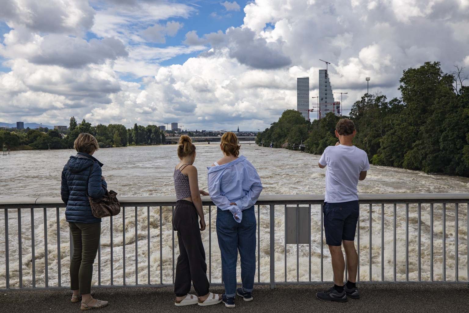 epa09348555 People look on from a bridge amid high water levels on the Rhein (Rhine River), as the Roche Towers stand in the background, in Basel, Switzerland, 16 July 2021. The high water mark was ex ...