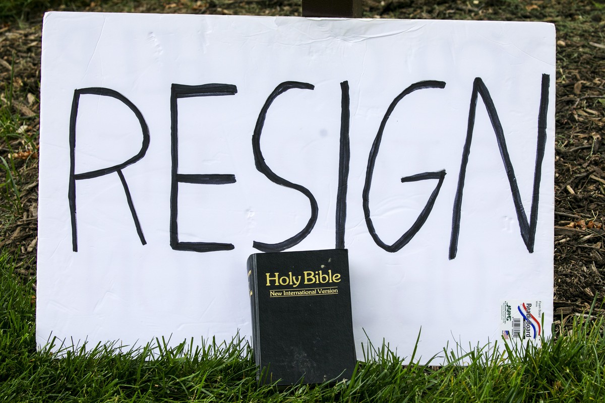 A sign says "resign" as a Bible is propped against it during a protest of the arrival of President Donald Trump to the Saint John Paul II National Shrine, Tuesday, June 2, 2020, in Washingto ...